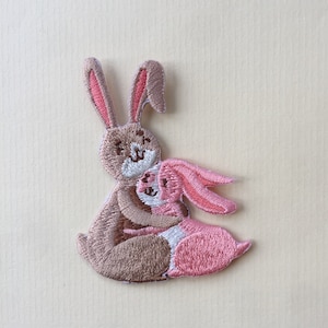Op de afbeelding: Gestikt patch met twee bruine en roze konijnen die elkaar knuffelen. Het grotere konijn is bruin met roze oren en het kleinere konijn is roze met bruine oren.