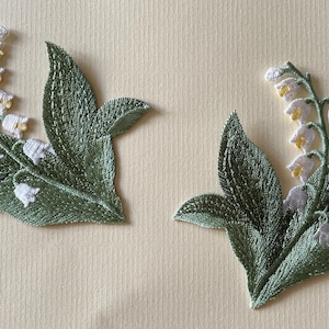 May include: Two embroidered lily of the valley floral patches. Each patch features white bell-shaped flowers with yellow centres and green leaves. The patches are on a light beige background.