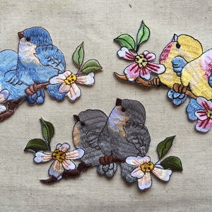 Puede incluir: Tres parches bordados para planchar que representan parejas de pájaros posados en ramas con flores rosas y blancas. Los pájaros son azules, amarillos y grises.