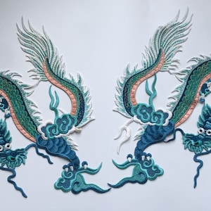 May include: Two embroidered iron-on patches featuring a blue and green dragon with white and peach accents. The dragons are facing each other with their wings spread out.