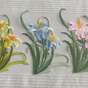 May include: Embroidered lily flower patches in yellow, blue, and pink. Each patch displays detailed stitching of the petals, green leaves, and stems. The patches are set against a light grey fabric background, showcasing the floral designs.