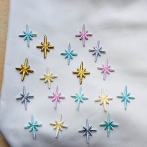 May include: A collection of 18 star-shaped iron-on patches in various colours, including gold, yellow, pink, blue, and teal. The patches are arranged in a grid pattern on a white fabric background.