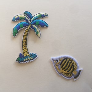 May include: Two embroidered iron-on patches, one of a palm tree with green, blue, and yellow colours, and the other of a yellow and black fish with white outlines.