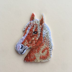 以下が含まれることがあります： 茶色と白の斑点模様の馬の頭が刺繍されたアイロン接着パッチ。馬は白いたてがみと茶色の鼻をしています。