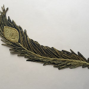 Könnte beinhalten: Ein gestickter Pfauenfeder-Patch in Schwarz und Gold. Die Feder ist mit goldfarbenem Garn verziert und hat einen schwarzen Umriss.