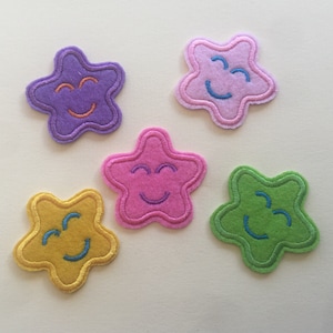 May include: Five felt star-shaped patches with smiling faces. The stars are purple, pink, yellow, green, and light pink. Each star has a different facial expression.