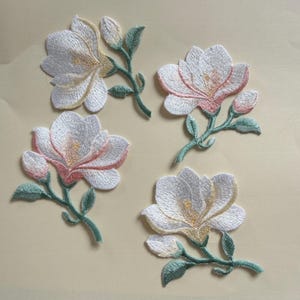 以下が含まれることがあります： 白のモクレンの花と黄とピンクのアクセントが特徴の、4つの刺繍された花のパッチ。各パッチには緑の茎と葉が含まれています。パッチは薄いベージュ色の背景にあります。
