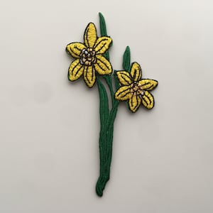 May include: A yellow embroidered patch of two daffodils with green stems and leaves.
