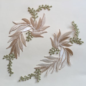 May include: Two beige and white embroidered floral appliques with green glitter accents. The appliques are arranged in a circular pattern on a light beige background.