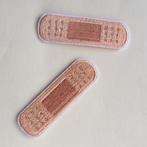 Könnte beinhalten: Zwei gestickte Stoff-Patches in Form von Pflastern. Die Patches sind beige mit brauner Mitte und weißen Nähten an den Rändern.