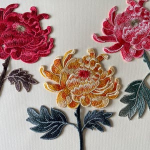 May include: Embroidered floral patches featuring chrysanthemum blooms in shades of red, orange, and yellow. Each flower has detailed stitching and is attached to a stem with leaves. The patches are ideal for adding a decorative touch to clothing or accessories.