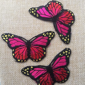 Pode incluir: Três patches bordados de borboletas rosa e vermelhas com contornos pretos e detalhes amarelos.