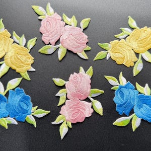 Op de afbeelding: Een verzameling van vijf geborduurde rozenpatches, bestaande uit twee roze rozenparen, twee gele rozenparen en één blauw rozenpaar. Elke bloemapplicatie bevat gedetailleerd stikwerk met lichtgroene bladeren en witte accenten op de bloemblaadjes.