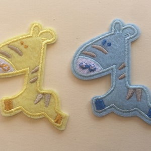 May include: Two embroidered giraffe patches. One is yellow with brown stripes and accents, the other is light blue with brown stripes and accents. Both have embroidered details on the face and feet. These patches are likely for clothing or crafts.