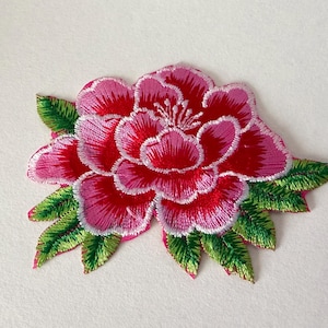 Puede incluir: Parche floral bordado que presenta una flor grande y estilizada con pétalos en capas. La flor es principalmente roja y rosa, con contornos blancos y un centro detallado. Hojas verdes con detalles blancos rodean la flor, creando un diseño decorativo.