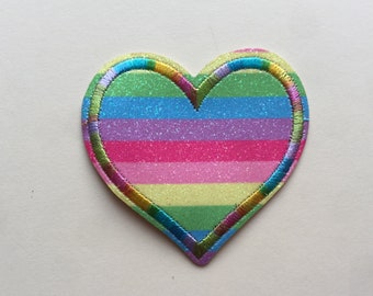 May include: A heart-shaped patch with a rainbow striped design. The patch is made of glitter fabric and has a colorful embroidered border.