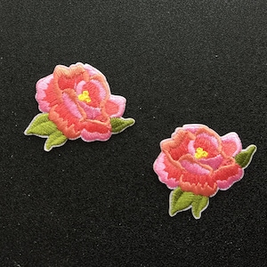 May include: Two pink embroidered fabric flowers with green leaves. The flowers have yellow centers.