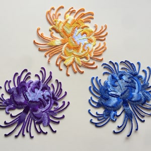 May include: Three embroidered iron-on patches in the shape of chrysanthemums. One is yellow, one is purple, and one is blue.
