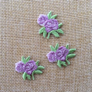 Könnte beinhalten: Drei gestickte Blumenapplikationen in Lila und Grün auf braunem Jutestoff.