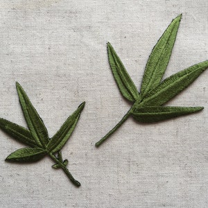 Puede incluir: Dos hojas de tela bordadas de color verde con forma puntiaguda y múltiples puntas. Las hojas están sobre un fondo de lino blanco.