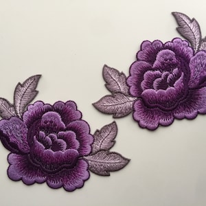 Può includere: Due applicazioni di fiori in tessuto ricamate di colore viola con foglie grigie. I fiori sono in stile peonia.