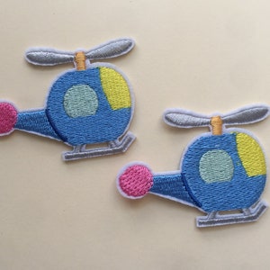 May include: Two blue embroidered iron-on patches of helicopters. Each helicopter has a yellow and light blue detail on the side, a pink circle on the front, and a silver propeller.