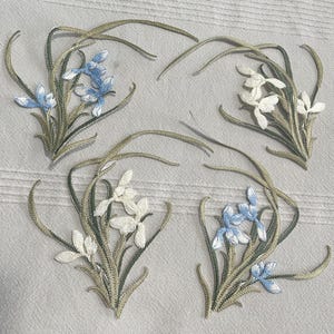 May include: Embroidered floral patches featuring iris flowers in white and light blue. The patches have green stems and leaves with a gold outline. The patches are arranged on a light gray fabric.