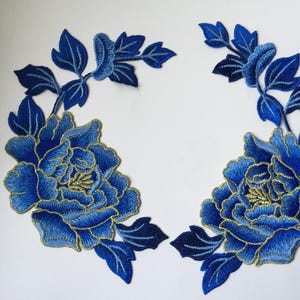 Op de afbeelding: Twee blauwe geborduurde stoffen applicaties met een bloemmotief en gouden accenten. Elke applicatie heeft een grote bloem met bladeren en stengels.
