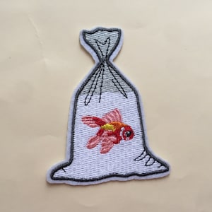 May include: Embroidered patch depicting a goldfish in a plastic bag. The fish is red and yellow, with detailed stitching. The bag is white with a grey outline and black drawstring. The patch has a white border.