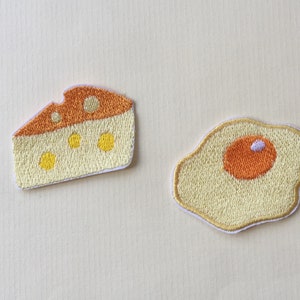 以下が含まれることがあります： 黄色とオレンジのステッチで刺繍されたチーズの形をしたアイロン接着パッチと、黄色とオレンジのステッチで刺繍された目玉焼きの形をしたアイロン接着パッチの2つです。