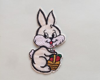 May include: Embroidered patch featuring a cartoon bunny holding a basket of fruit. The bunny is white with black outlines and details, including a red tongue. The basket is gold with red and green fruit.