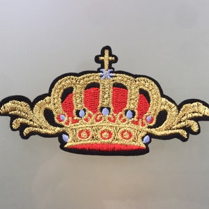 Puede incluir: Un parche bordado de una corona dorada con detalles rojos y azules. La corona tiene una punta puntiaguda y un diseño decorativo.