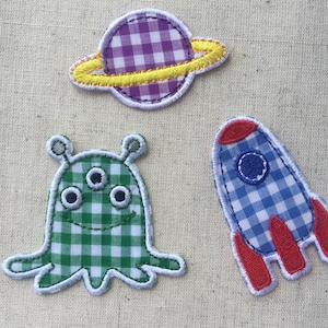 Puede incluir: Tres aplicaciones de fieltro con temática espacial. Un planeta morado y amarillo, un extraterrestre verde con tres ojos y un cohete azul y rojo.