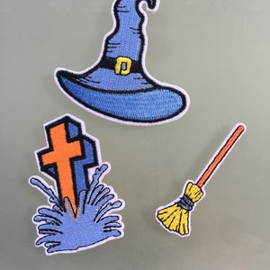 May include: Three embroidered patches: a blue witch's hat with a black band and gold buckle, a tombstone with an orange cross and blue lightning, and a broom with a yellow straw head and orange handle.