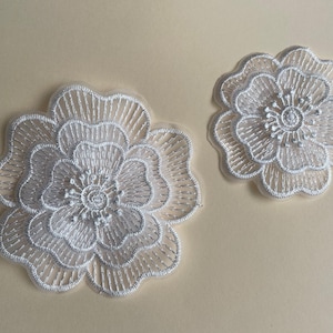 Può includere: Due applicazioni di fiori in pizzo bianco. L'applicazione più grande ha un diametro di circa 10 cm e quella più piccola di circa 5 cm.