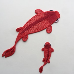 以下が含まれることがあります： 赤い刺繍の魚のパッチが2つ。大きな魚は右に泳いでいて、小さな魚は上に泳いでいます。両方の魚は黒い目を持っています。