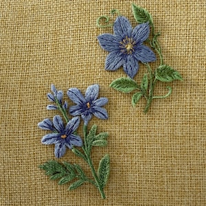 Könnte beinhalten: Zwei gestickte Blumenaufnäher mit blauen Blüten, gelben Mitten und grünen Blättern. Ein Aufnäher zeigt eine einzelne große Blüte mit geschwungenem Stiel, während der andere eine Gruppe aus zwei kleineren Blüten an einem beblätterten Stiel darstellt.