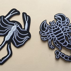 May include: Two embroidered iron-on patches featuring scorpions. The scorpions are outlined in white and filled in with black. One scorpion is curled up with its tail raised, while the other scorpion is standing with its tail down.