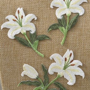 May include: Three embroidered lily patches featuring white petals with pale yellow centres, green stems, and dark brown stamens. Each patch displays detailed stitching of the flower blossoms and pointed green leaves, creating a textured appearance.