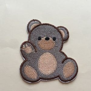 Könnte beinhalten: Ein gestickter Teddybär-Patch in Grau mit braunen Akzenten. Der Bär sitzt mit erhobenem linken Pfote und hat einen freundlichen Ausdruck.