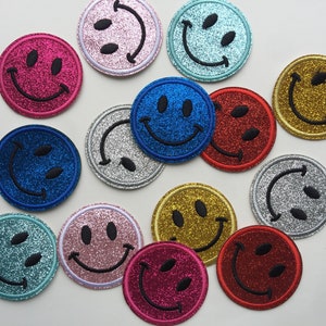 May include: Assortment of 12 iron-on patches featuring smiley faces in various colours and glitter finishes. The patches are approximately 5 cm in diameter.