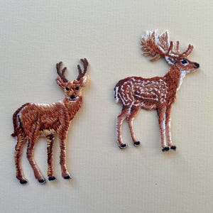 May include: Two embroidered deer patches. One deer is standing with antlers, the other has a more elaborate antler design. Both patches are brown with white accents and black hooves, set against a neutral background.