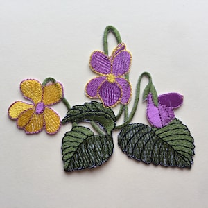 Puede incluir: Un parche bordado con un grupo de flores moradas y amarillas con hojas verdes. Las flores están en un estilo delicado y vintage.