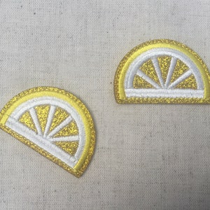 Peut inclure: Deux patchs en feutre en forme de tranches de citron jaunes et blanches. Les tranches de citron sont des demi-cercles avec un contour blanc et un centre jaune avec des lignes blanches rayonnant du centre.