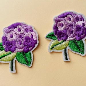Op de afbeelding: Twee geborduurde bloemenpatches in paars en wit met groene bladeren. De bloemen zijn in een tros gerangschikt en hebben een 3D-effect.