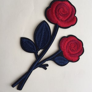 Könnte beinhalten: Gestickter Aufnäher mit zwei roten Rosen mit schwarzen Konturen und marineblauen Stielen und Blättern. Die Rosen haben detaillierte Stickereien, und die Blätter haben ein strukturiertes Aussehen. Der Aufnäher ist für dekorative Zwecke gedacht, z. B. für Kleidung oder Accessoires.