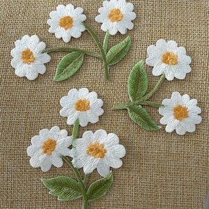 May include: Three embroidered daisy patches featuring white petals and yellow centres. Each patch includes green stems and leaves. The embroidery is detailed with visible stitching patterns, showcasing the floral design in a clear, flat arrangement.