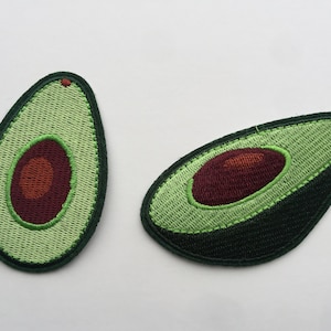 Puede incluir: Dos parches bordados en forma de aguacate verde con huesos marrones. Los parches están sobre un fondo blanco.
