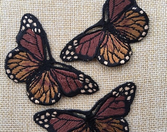 May include: Three embroidered butterfly patches in shades of brown and burgundy. The butterflies have black outlines and white spots.