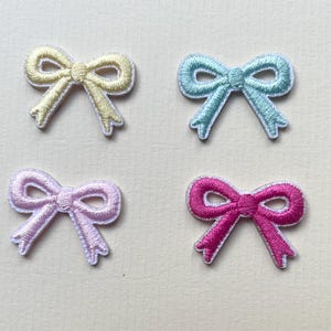 Puede incluir: Cuatro parches de lazo de tela bordados en diferentes colores: amarillo, azul claro, lavanda y rosa. Todos los lazos son del mismo tamaño y forma.